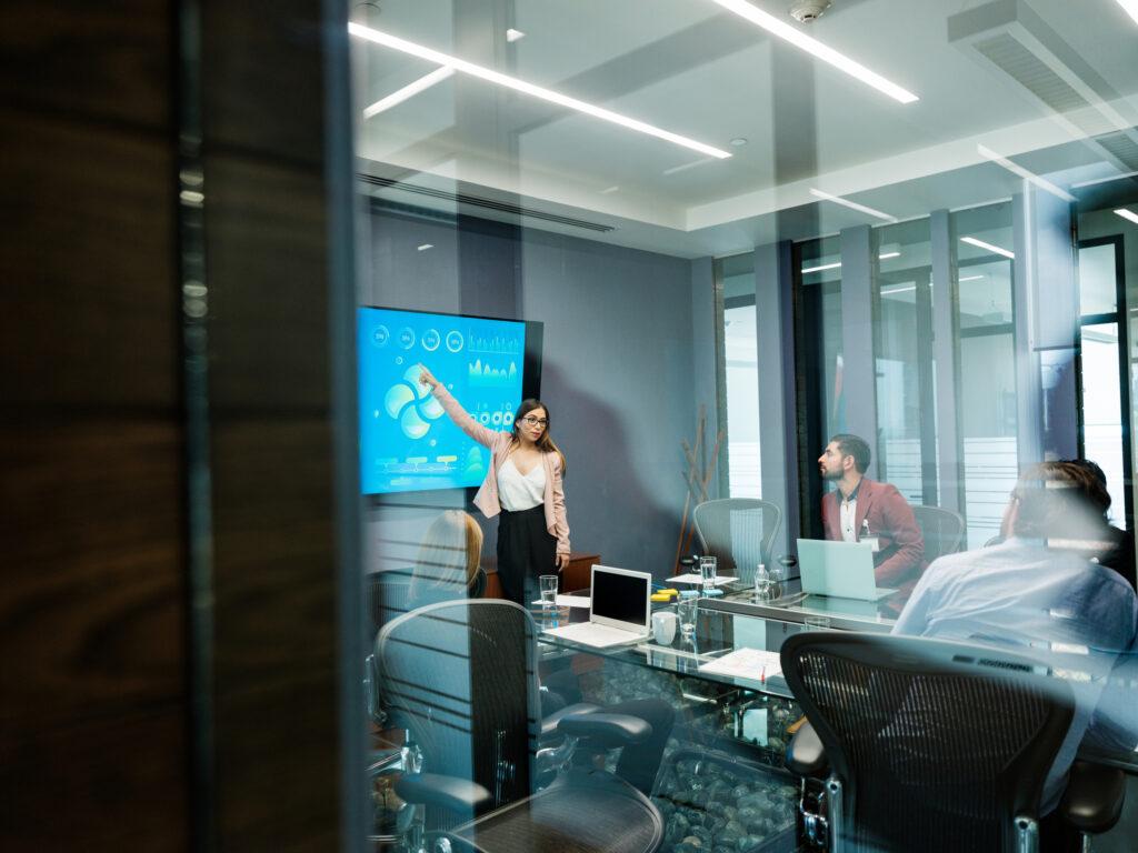 Riunione in sala meeting videoconferenza con display interattivo e partecipanti attorno al tavolo.