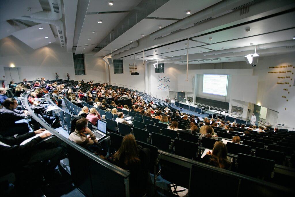 Lezione in auditorium universitario con grande schermo e videoproiettore durante presentazione accademica.