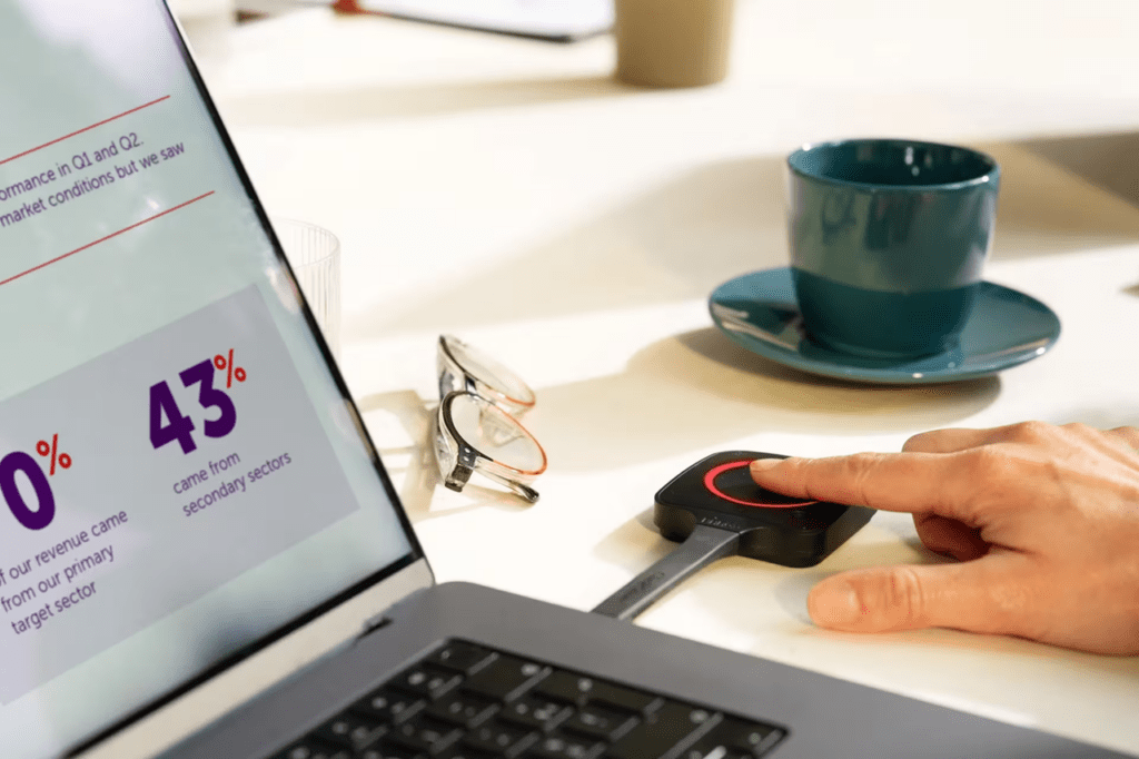 Mano che utilizza un dispositivo Barco Clickshare per avviare una videoconferenza su laptop con slide di dati.